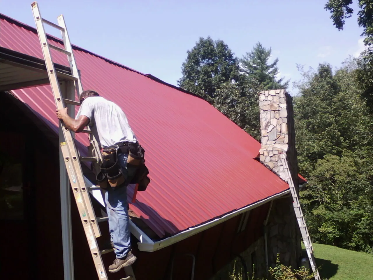 Roofer on a ladder beside a red metal roof and stone chimney — Soffit & Fascia Repair by Roofing Brothers in Oakland Mills